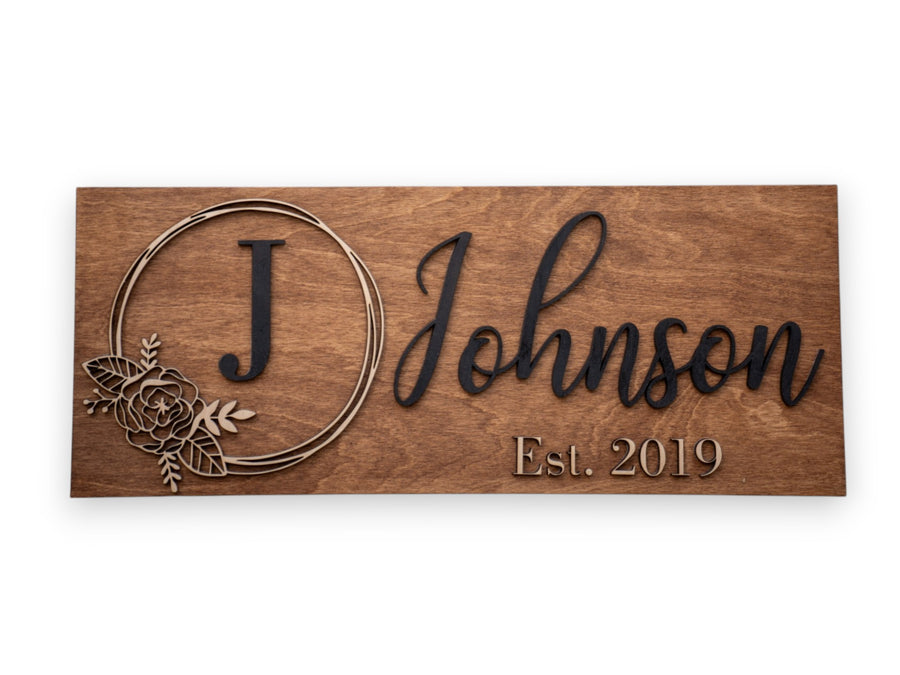 Walnut wood sign with a carved floral wreath around the letter J, reading Johnson, Est. 2019.