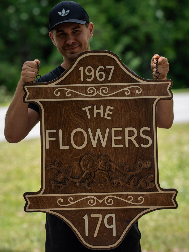 Shield-shaped dark walnut family sign reading 1967, The Flowers, 179, with an engraved rose and vine band and cream scroll borders