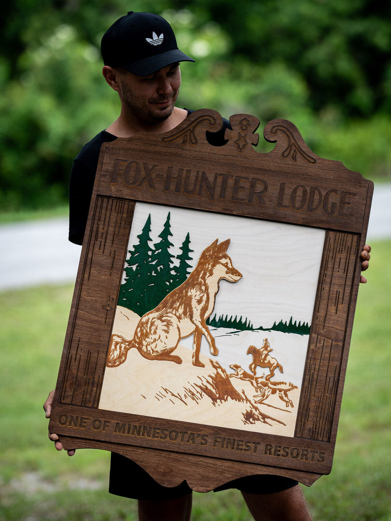 Large dark walnut resort sign with a carved scrolled crown and an engraved fox on a snowbank with pines and a distant huntsman