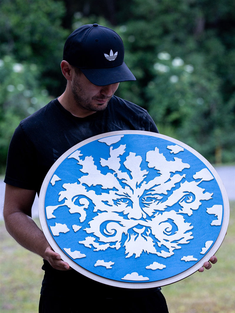 Round sky blue carved wood wall panel with swirling white cut-out oak foliage forming a bearded Green Man face