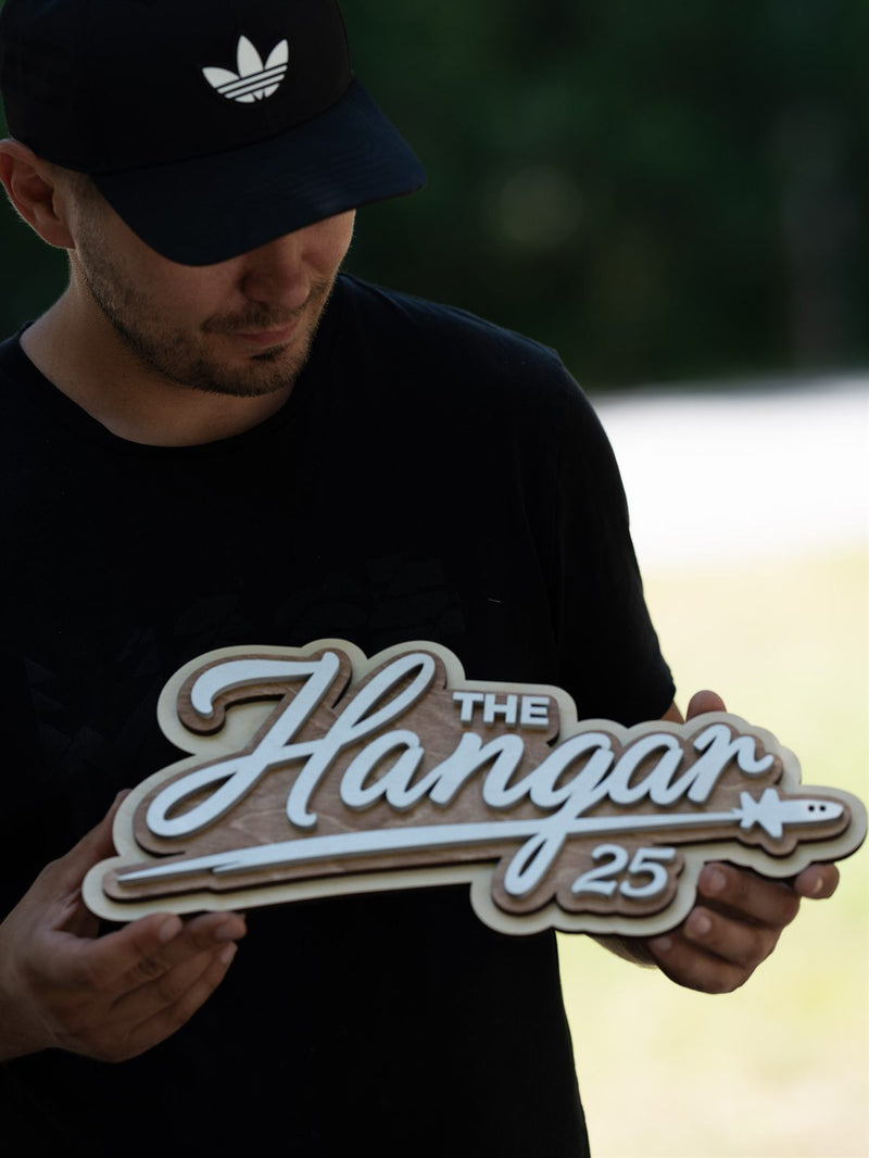 Long cut-out script wood sign reading The Hangar 25 with a swoosh underline ending in a small aeroplane silhouette