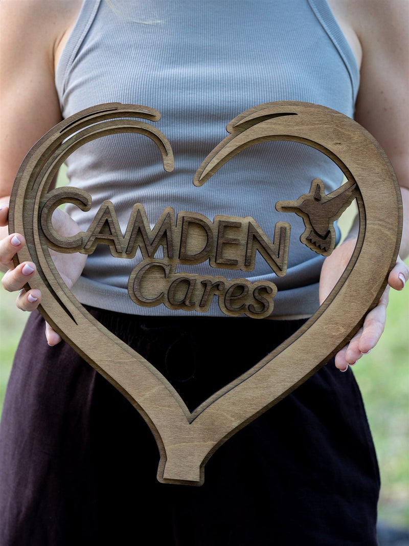 Open heart-shaped cut-out wood sign in natural stain reading Camden Cares with a hummingbird cut into the right lobe