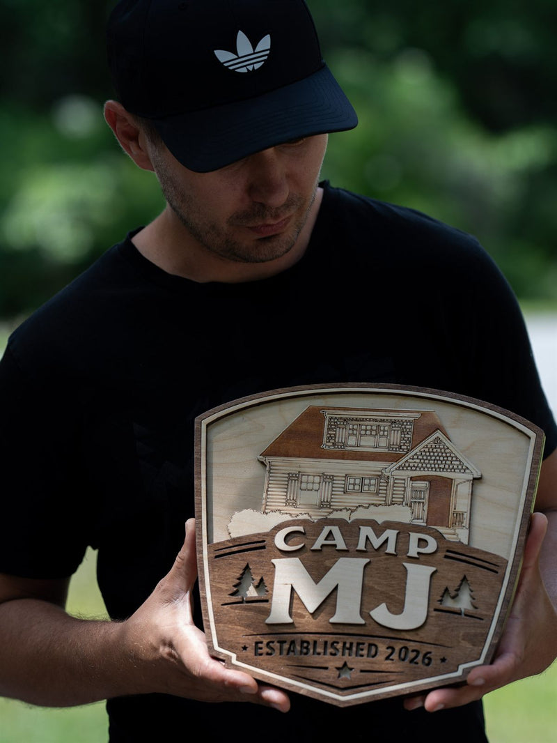 Shield-shaped wood cabin sign for Camp MJ with a finely engraved craftsman-style house in birch above dark walnut lettering