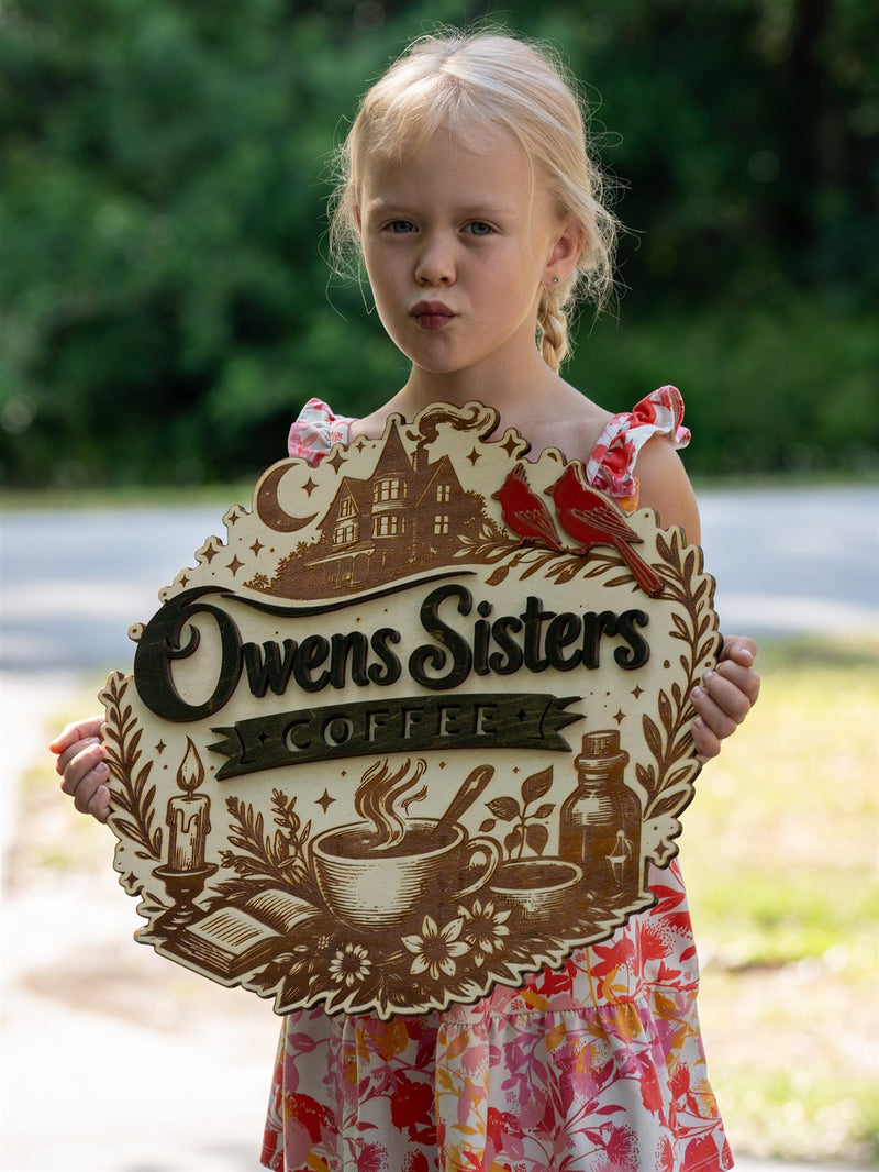 Cream scalloped wood coffee shop sign for Owens Sisters Coffee, engraved with a Victorian house, crescent moon, cardinals and a laurel border