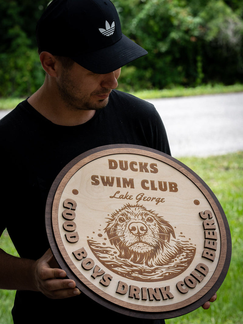 Round natural birch sign with a walnut outer ring, engraved with a wet shaggy dog swimming, reading Ducks Swim Club, Lake George
