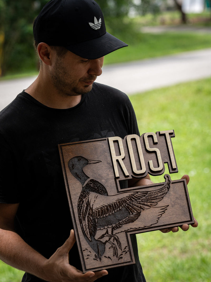 Rectangular dark walnut plaque deeply engraved with a loon landing on water, with pale birch block letters spelling Rost breaking over the corner