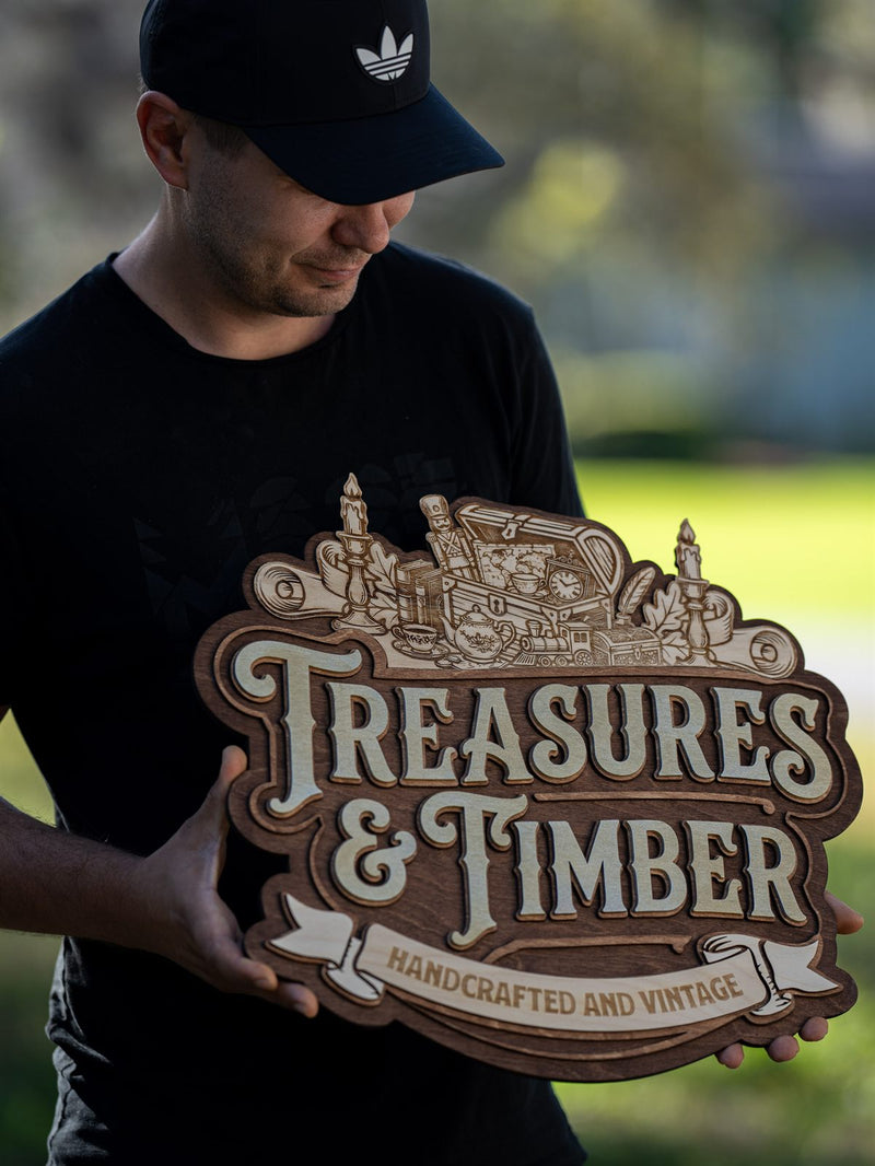 Engraved wood shop sign for Treasures and Timber with a still life of candlesticks, books, a clock and a toy train above ornate Victorian lettering