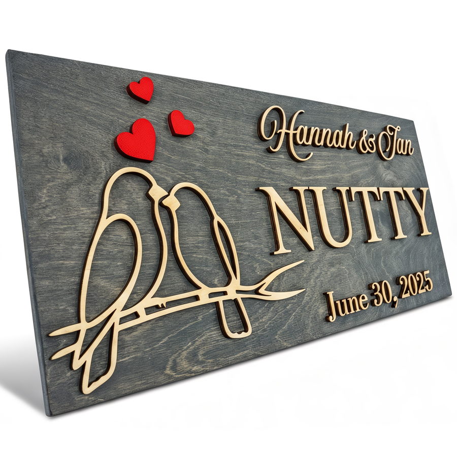 Grey wood sign with two carved lovebirds on a branch and red hearts, reading Hannah & Ian, NUTTY, June 30, 2025.