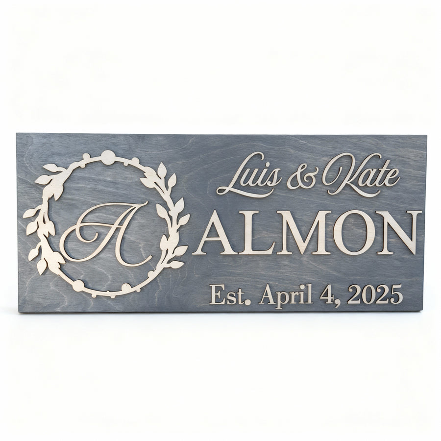 Slate grey wood sign with a carved laurel wreath around the letter A, reading Luis & Kate, ALMON, Est. April 4, 2025.