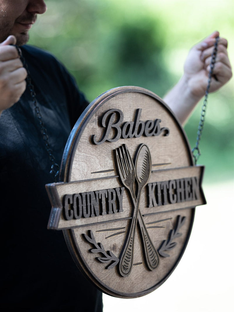 Oval wood restaurant sign hanging from two black chains with a raised script name, engraved fork and spoon and a ribbon banner