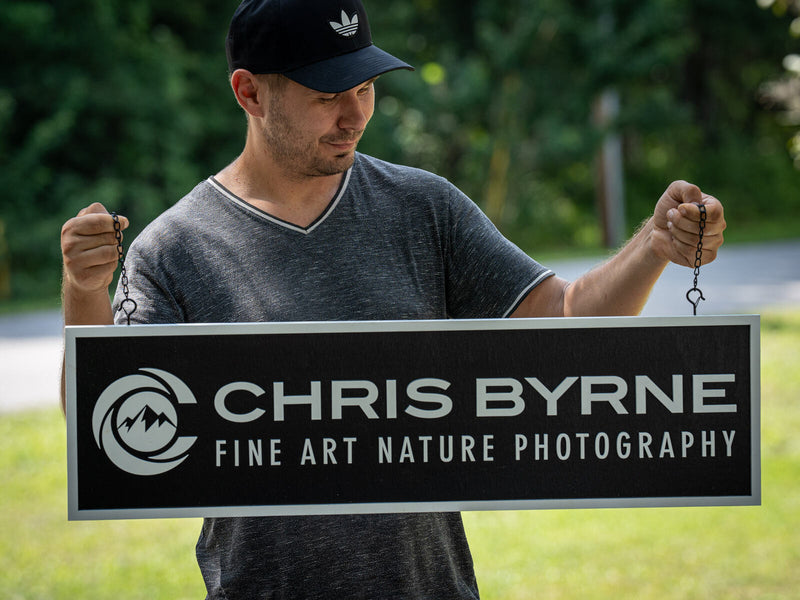 Long low charcoal grey wood studio sign hanging from a hook with white lettering reading Chris Byrne, Fine Art Nature Photography