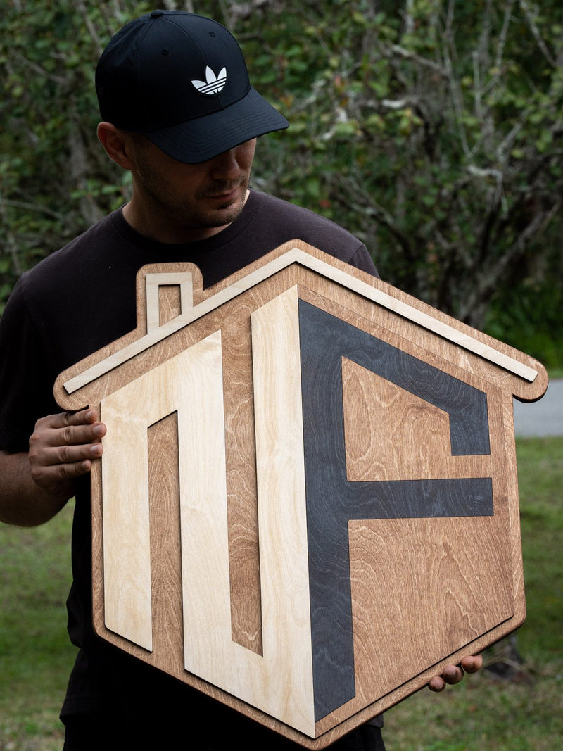 House-shaped wood logo sign in mid-brown stain with two interlocking angular letterforms in birch and charcoal