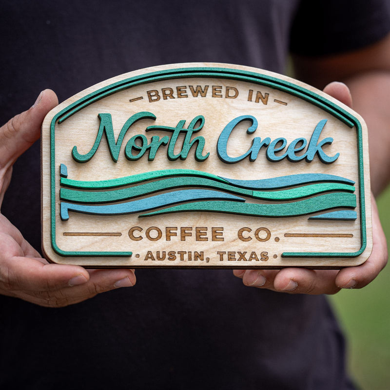 Close-up of a layered birch coffee company sign for North Creek Coffee Co, Austin Texas, with stacked teal and blue wave layers