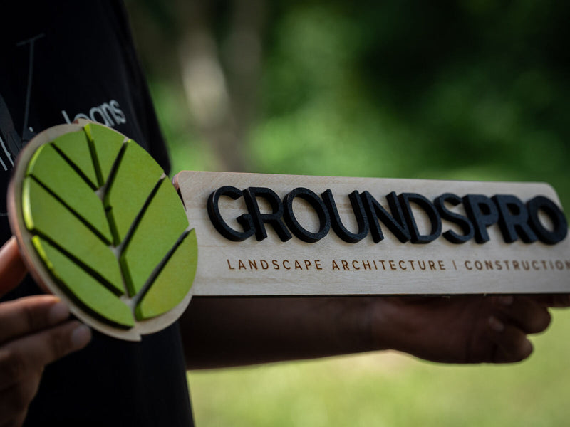 Extreme close-up along a long pale birch sign with thick black raised letters reading GroundsPro and a bright green layered leaf disc