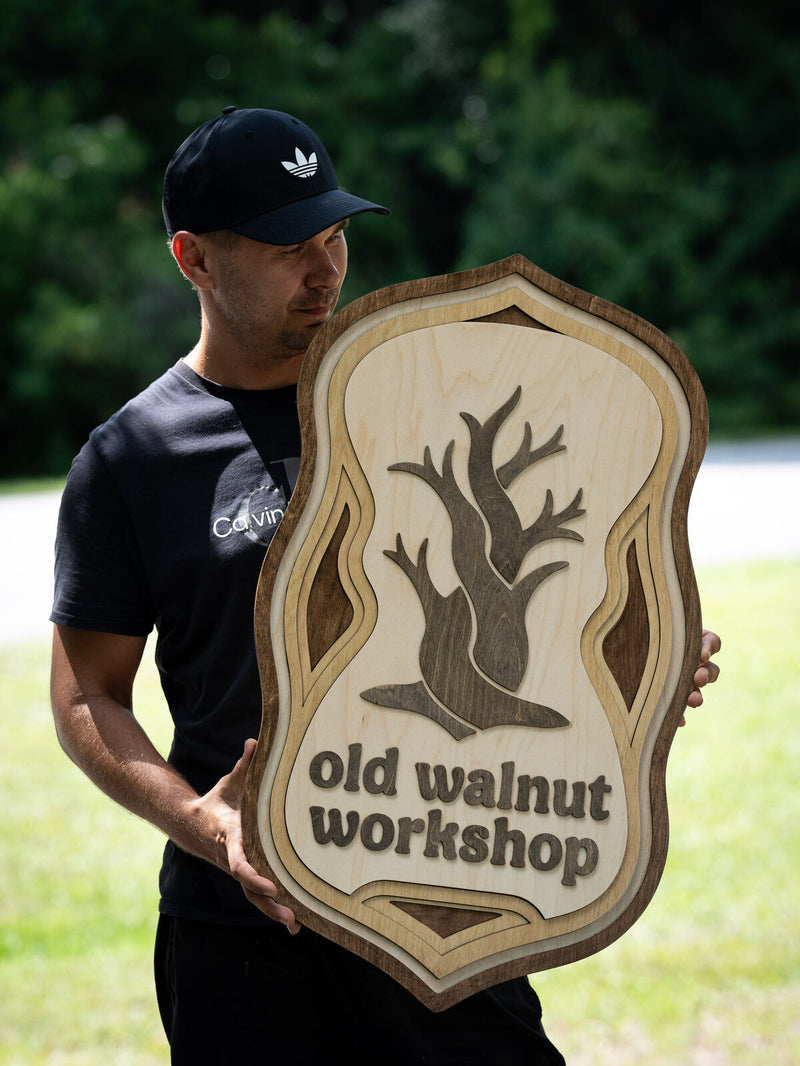 Tall ornate pale birch shield sign with layered scalloped borders and a bare walnut tree with spreading branches and roots at the centre