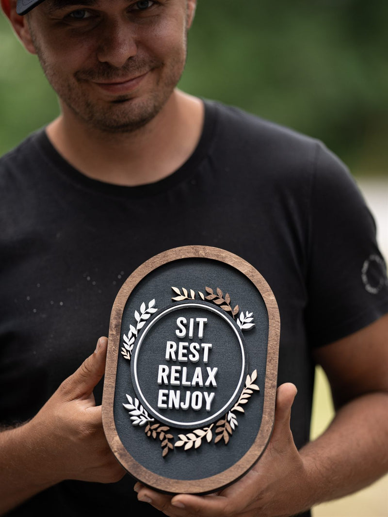 Small oval wood decor plaque with a mid-brown frame around a charcoal face reading Sit Rest Relax Enjoy with cream laurel sprigs