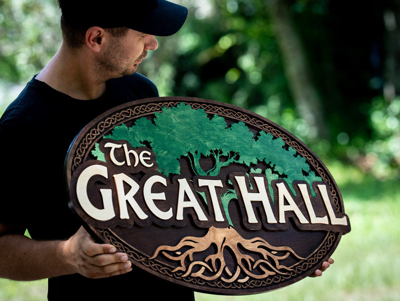 Wide oval wood sign with a scalloped dark walnut rim and a layered green oak tree with a broad canopy and exposed tan roots