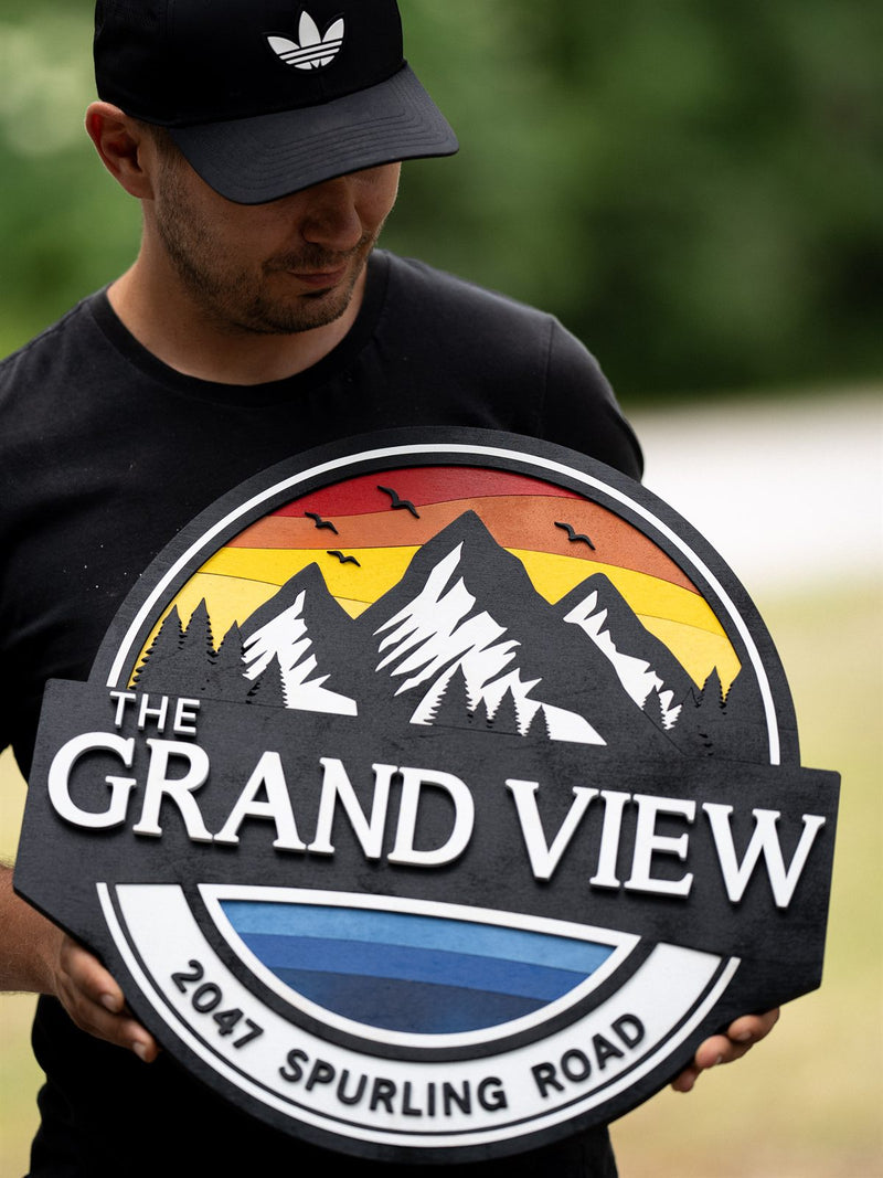 Round charcoal wood address sign with a retro red, orange and yellow sunset behind white-capped mountains, reading The Grand View, 2047 Spurling Road
