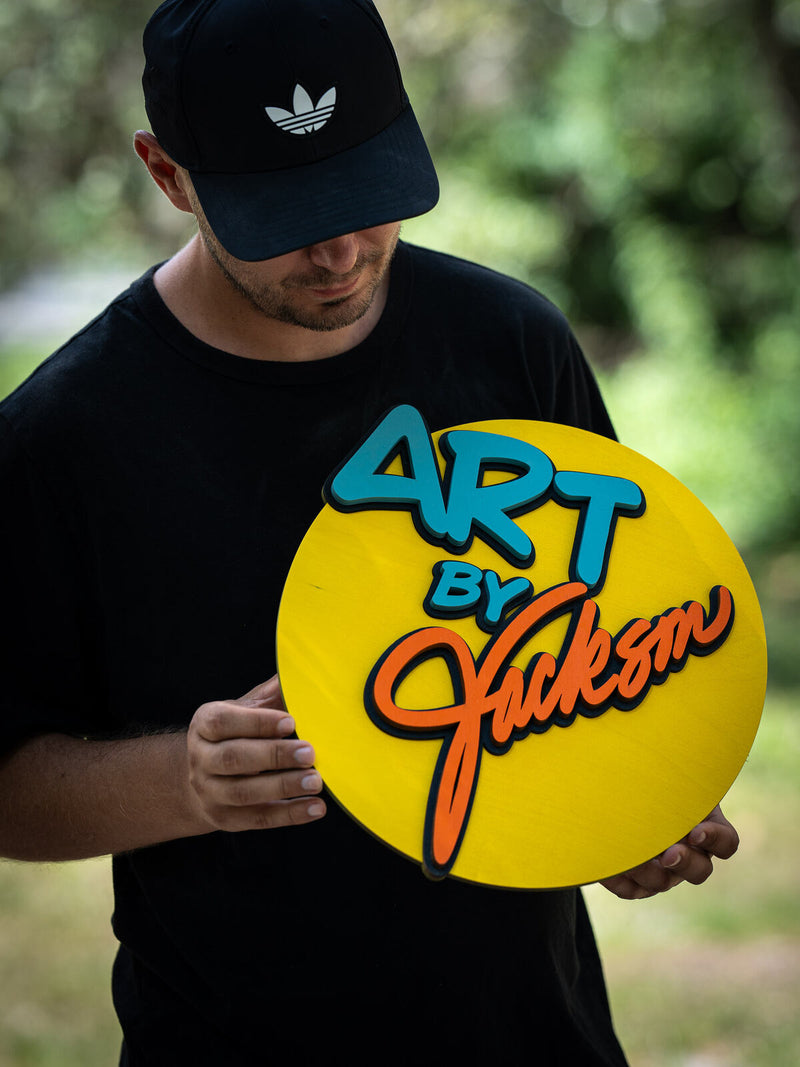 Bright yellow round wood studio sign with chunky teal blue lettering and an orange brush script name reading Art by Jackson