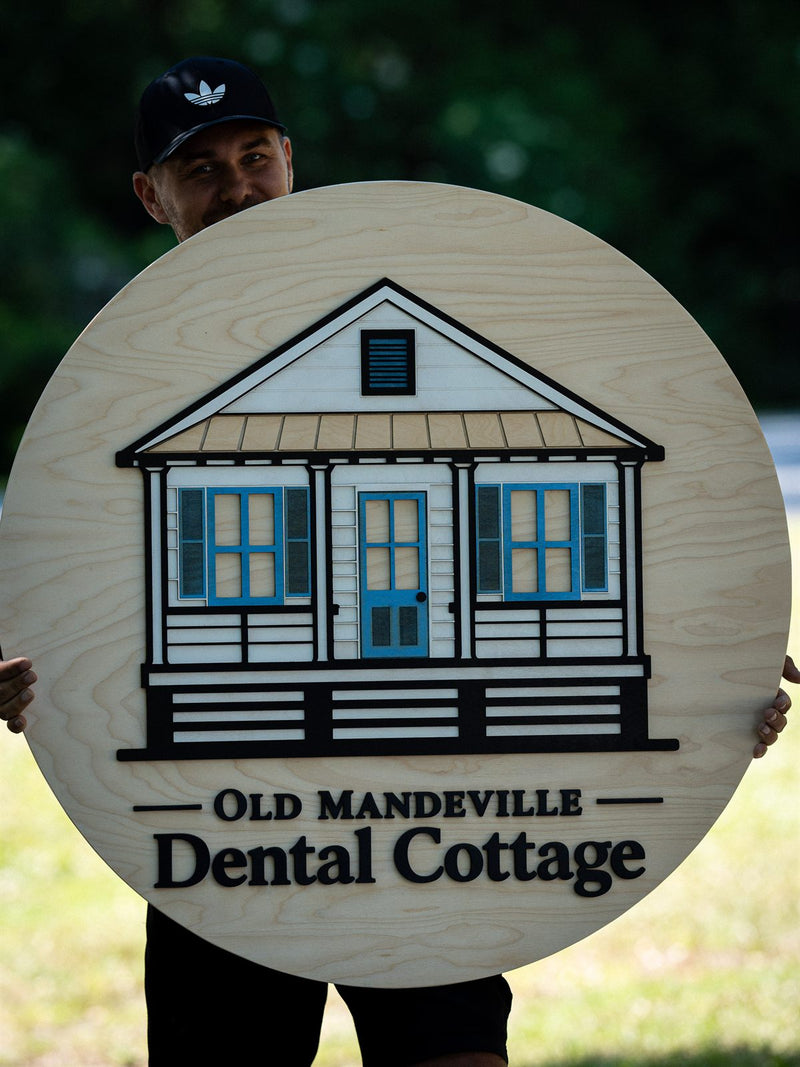 Large round pale birch dental office sign showing a layered Louisiana raised cottage with a teal door, reading Old Mandeville Dental Cottage