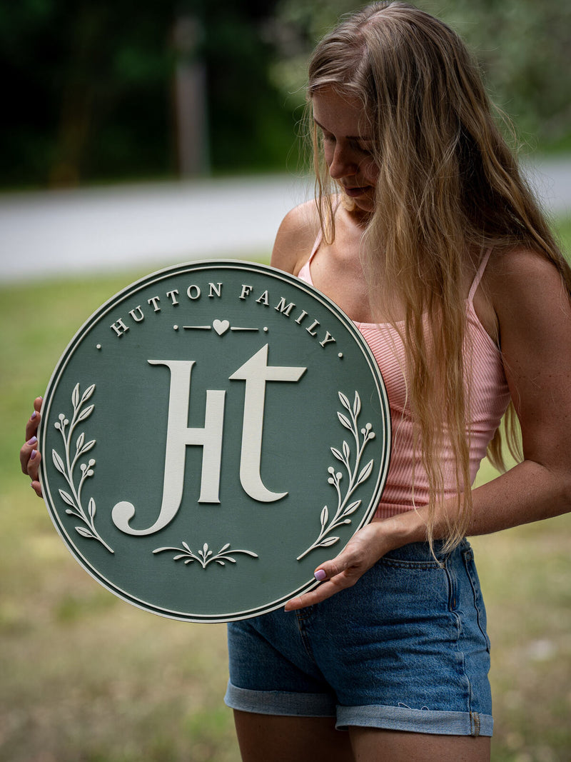 Large sage green round wood family sign with a tall cream Ht ligature monogram between two olive leaf sprigs