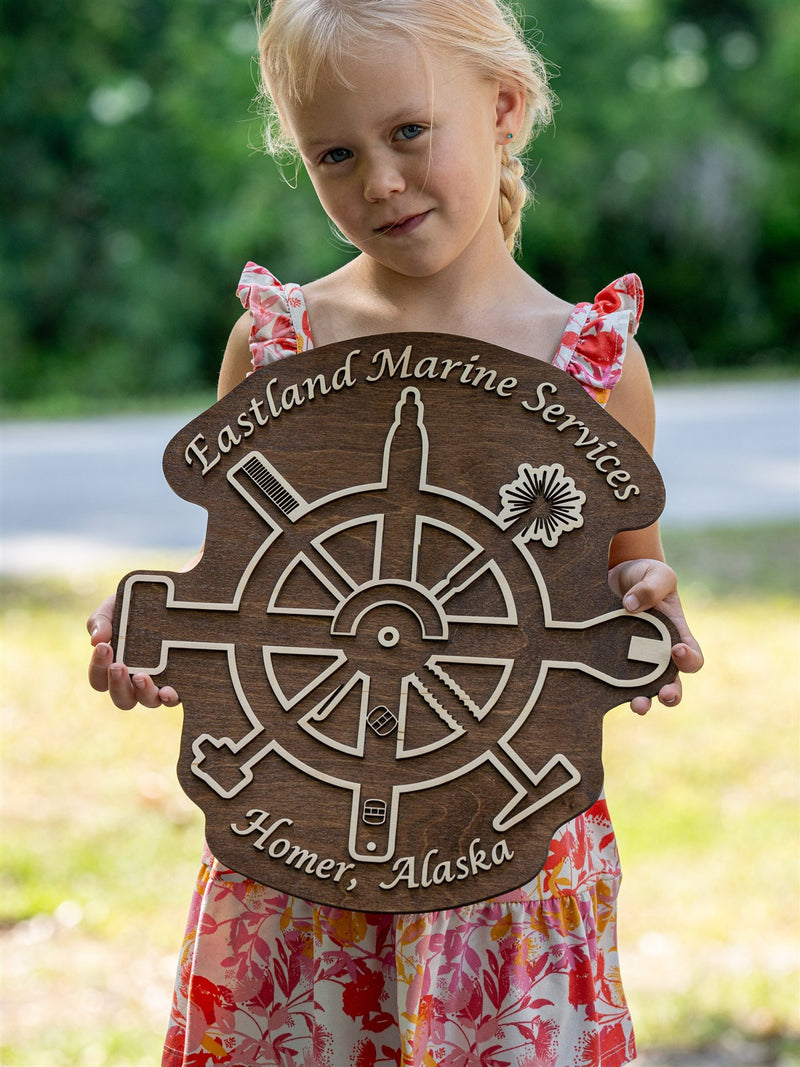 Round dark brown wood logo sign for Eastland Marine Services, Homer Alaska, engraved with a ship's wheel, anchor, oar and compass rose