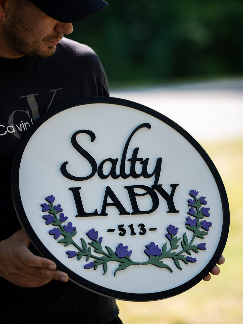 Round near-white wood name sign reading Salty Lady with a spray of purple lavender flowers on green stems