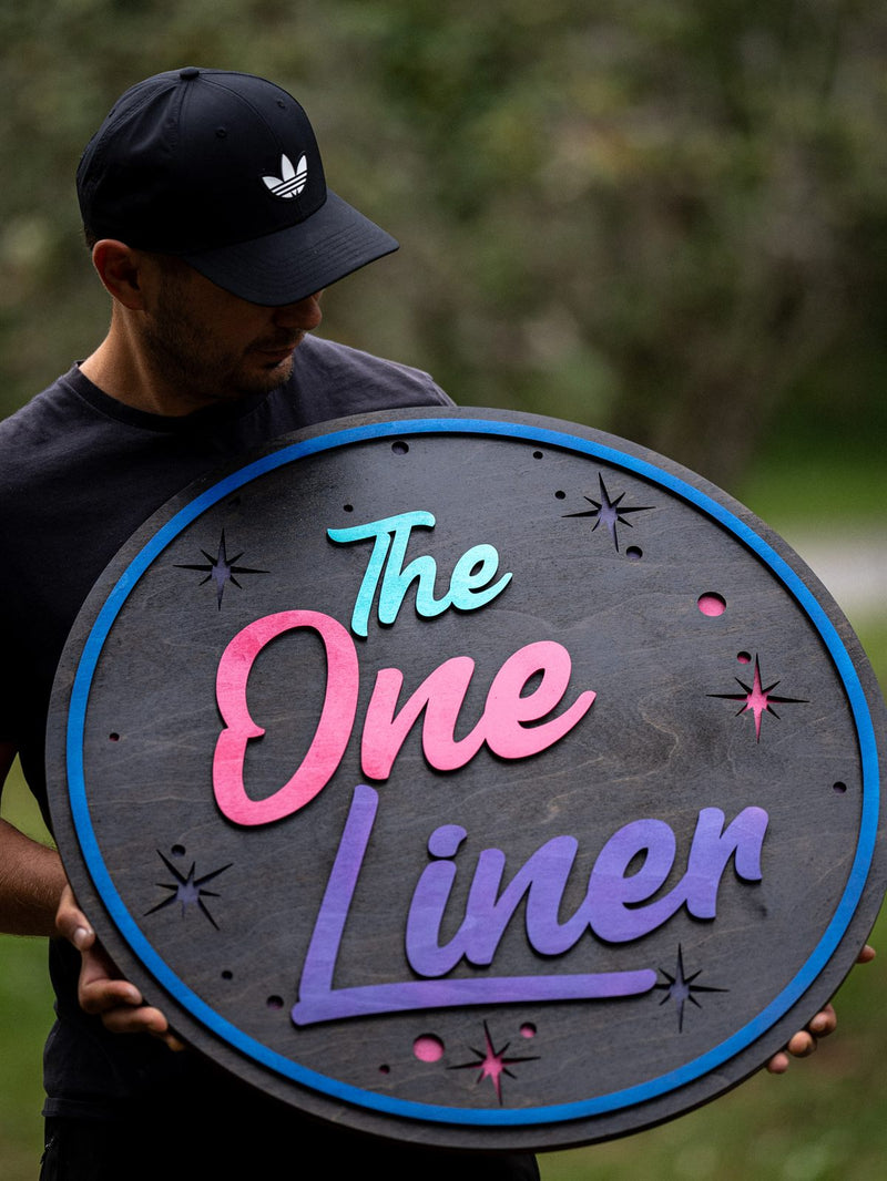 Round charcoal-stained wood sign with layered neon-style pink, mint and violet lettering reading The One Liner