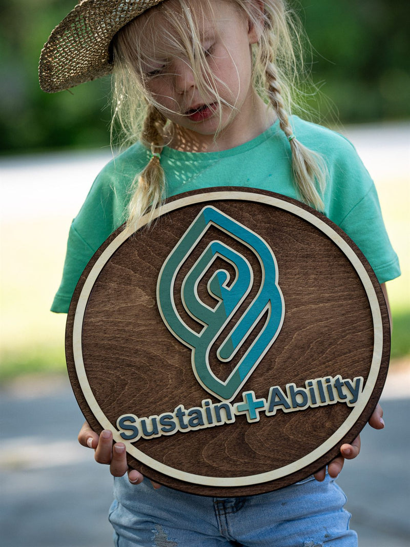 Round dark walnut office logo sign with a teal interlocking leaf monogram reading Sustain plus Ability