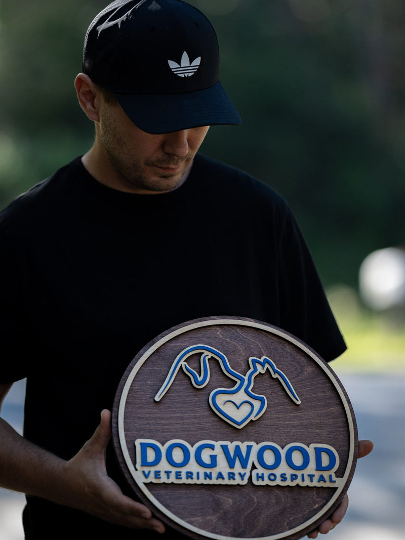 Round mid-brown wood veterinary sign with a blue and cream line drawing of a dog and cat sharing a heart, reading Dogwood Veterinary Hospital