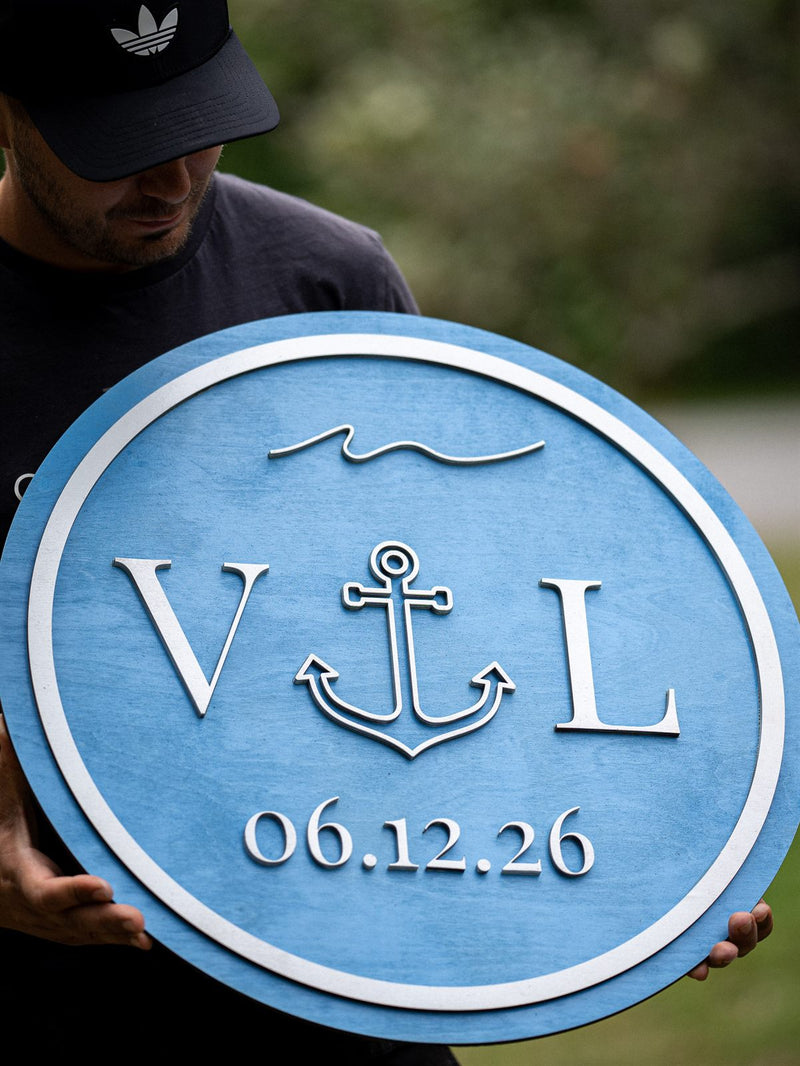 Round mid-blue stained wood wedding sign with white initials flanking an anchor and the date 06.12.26