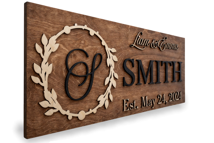 Walnut-stained wood sign with a carved laurel wreath around a monogram S, reading Liam & Emma, SMITH, Est. May 24, 2024.