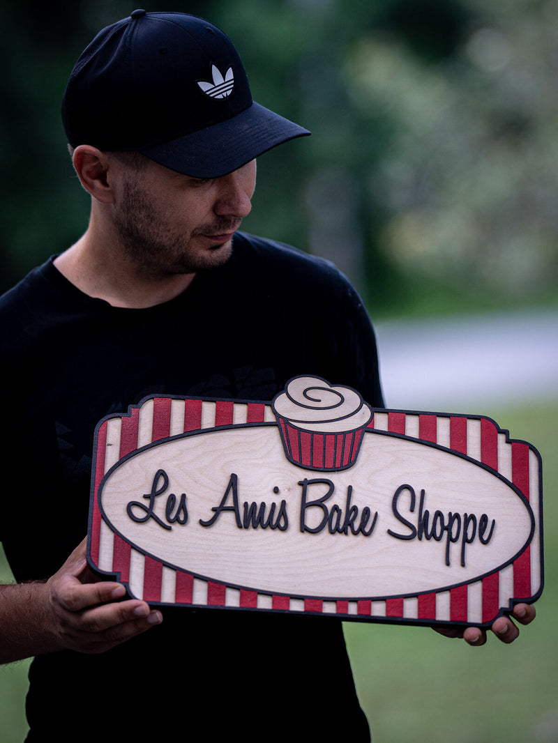 Rectangular red and white striped wood bakery sign with a cream oval carrying black script, and a striped cupcake breaking over the top edge
