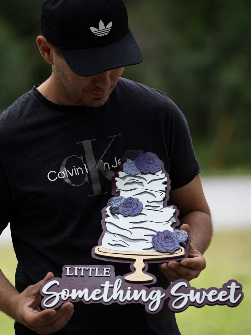 Wood bakery sign for Little Something Sweet with a layered three-tier ruffled wedding cake and purple roses on a plum backer