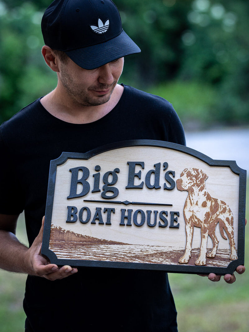 Rectangular pale birch boat house sign with a charcoal border, navy script lettering, an engraved dock scene and a layered harlequin Great Dane