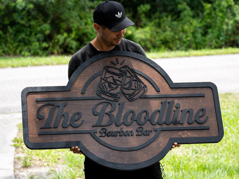 Wide walnut wood bar sign with a near-black raised border and two clinking rocks glasses outlined above black script reading The Bloodline Bourbon Bar