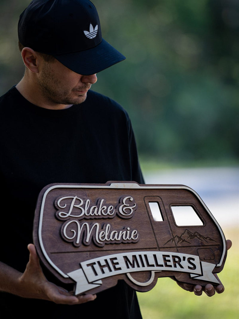 Wood sign cut in the silhouette of a vintage travel trailer, dark walnut with cream script names and an engraved mountain scene