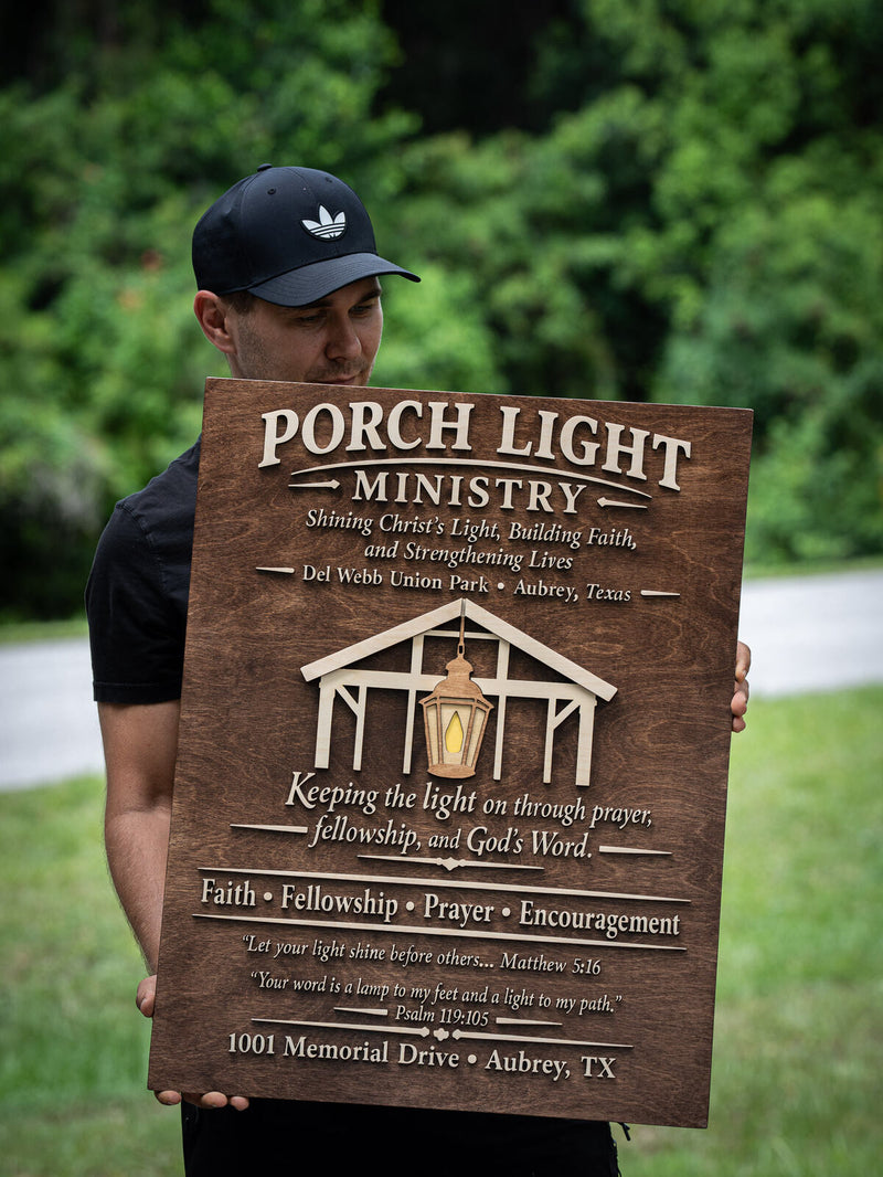 Tall rectangular walnut board covered in cream lettering with a layered pavilion frame and a hanging lantern, reading Porch Light Ministry