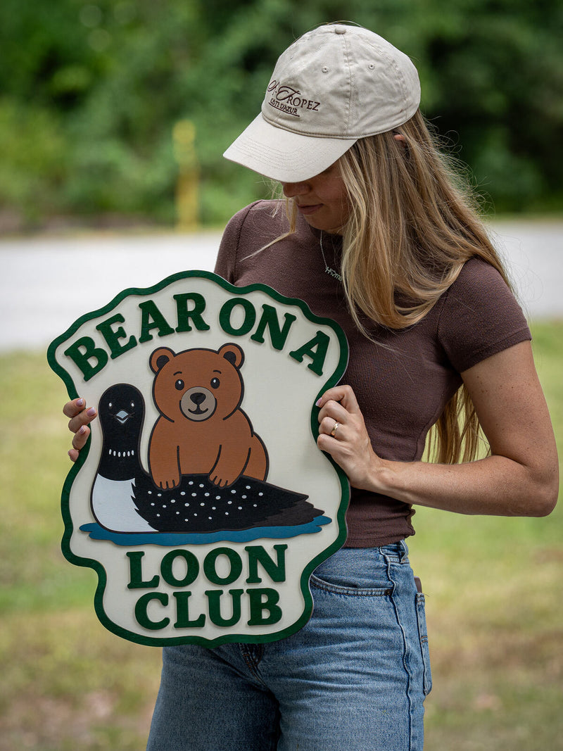 Cloud-shaped cream wood club sign with a dark green wavy border showing a cartoon bear cub riding a loon on blue water