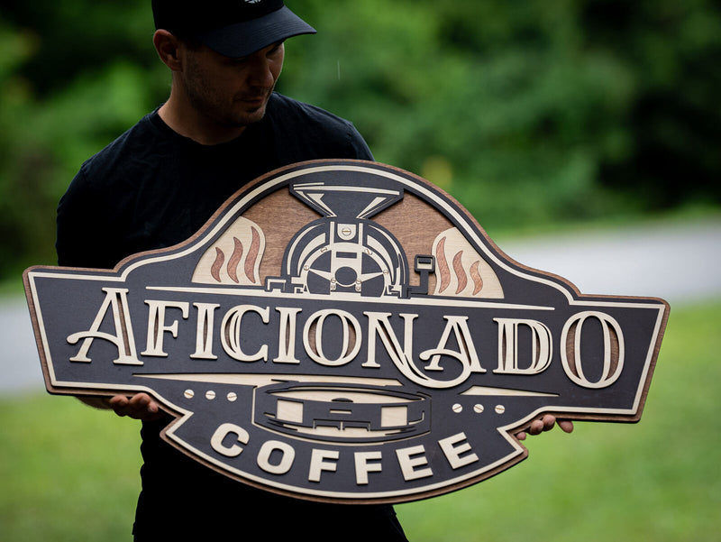Charcoal black shaped wood badge sign for Aficionado Coffee with a cream and tan drum coffee roaster and flame licks at the top