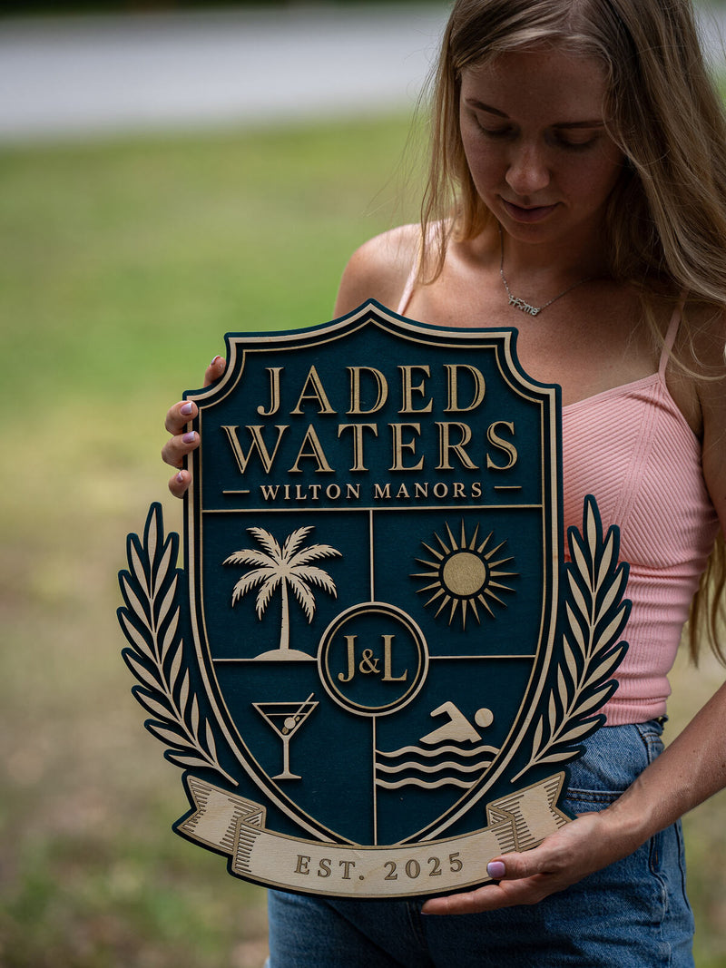 Tall deep teal shield-shaped wood crest for Jaded Waters, Wilton Manors, quartered with a palm tree, sunburst, martini glass and swimmer