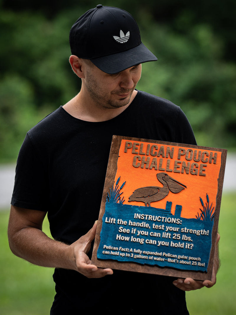 Square walnut-framed exhibit panel, orange top half with a brown pelican silhouette and reeds over a teal water panel of instruction text