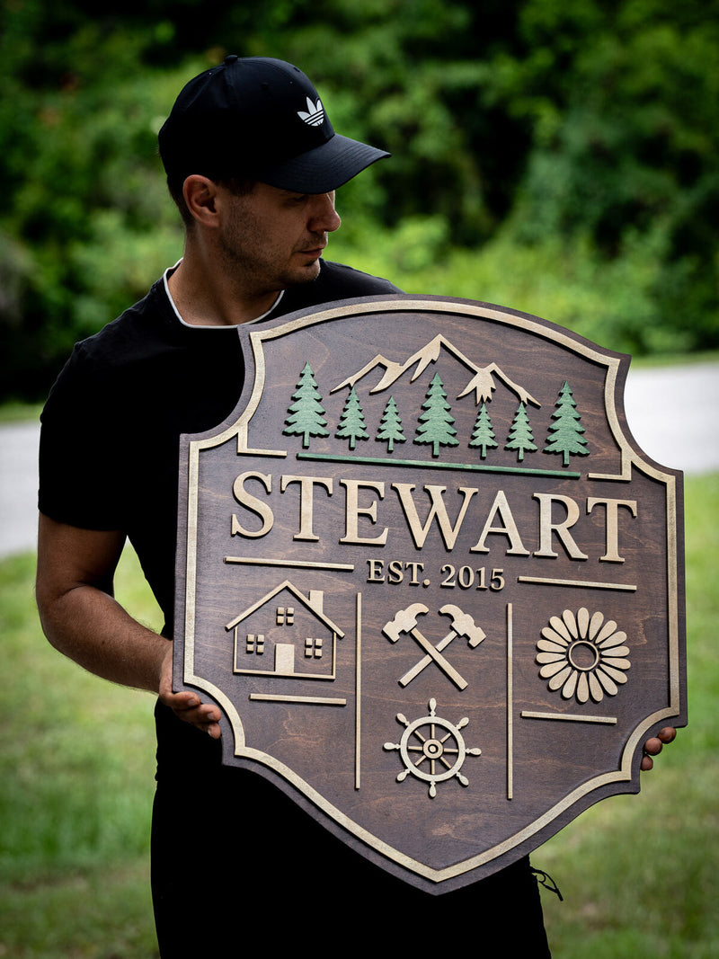 Walnut shield-shaped family sign with a stepped birch inner border, layered pines and mountains above the name Stewart, established 2015
