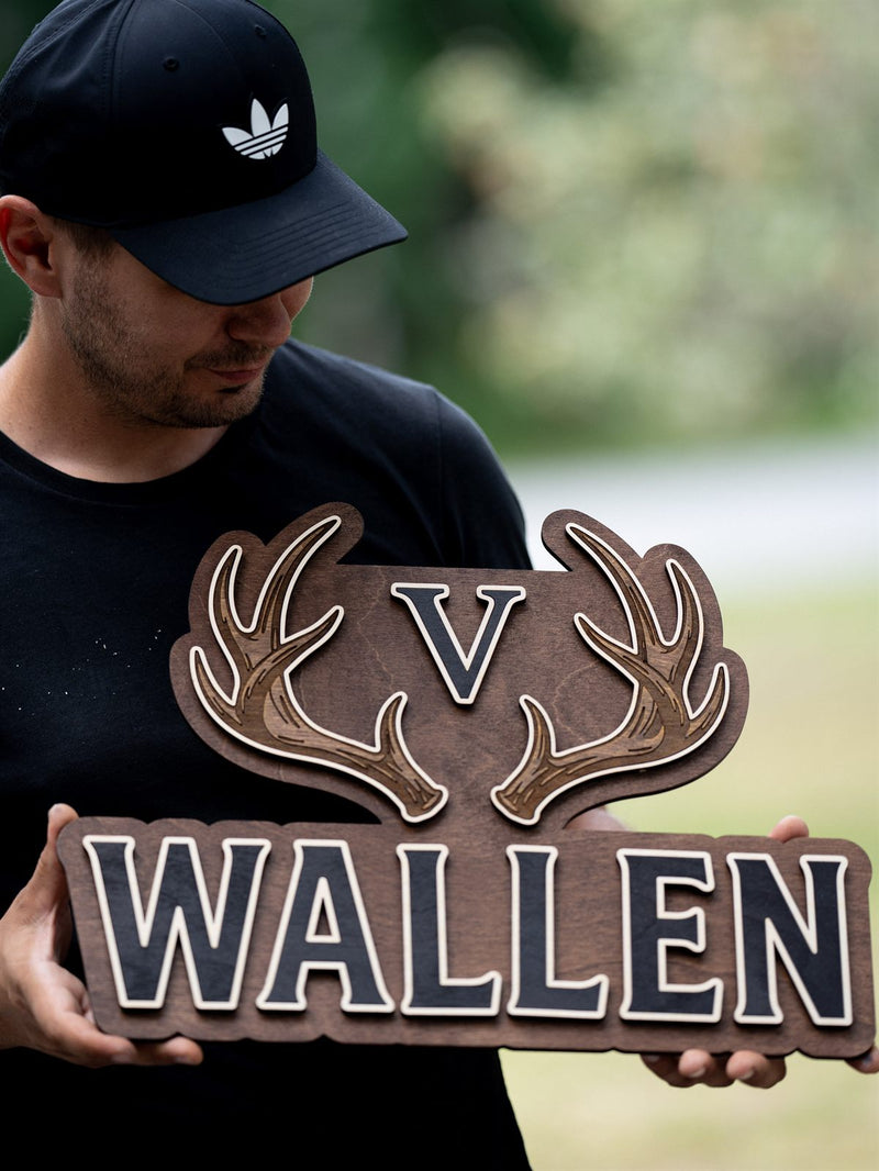 Wide warm brown wood family name sign shaped around a pair of layered antlers with a charcoal monogram between them