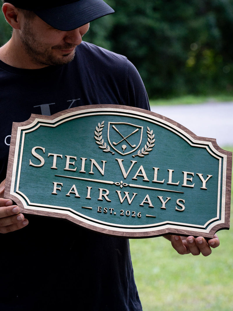 Clubhouse-shaped dark green wood golf sign with a walnut frame, a shield with crossed golf clubs, a tee'd ball and laurel sprays