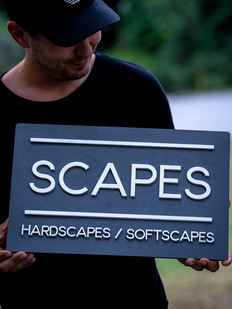 Plain slate-grey rectangular wood office sign with crisp white raised lettering reading Scapes, hardscapes, softscapes