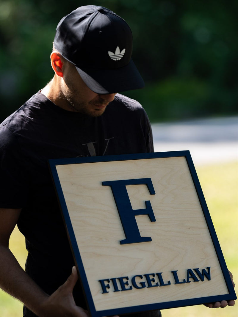 Square pale birch law firm lobby sign inside a navy border frame with a large navy serif F and the words Fiegel Law
