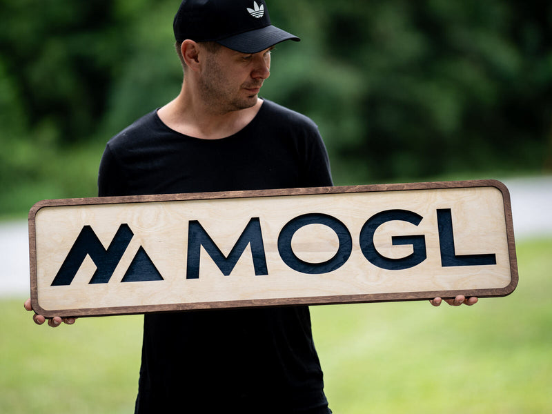Long low birch panel with rounded corners and a thin walnut edge, a navy triangular glyph beside heavy navy capitals reading MOGL