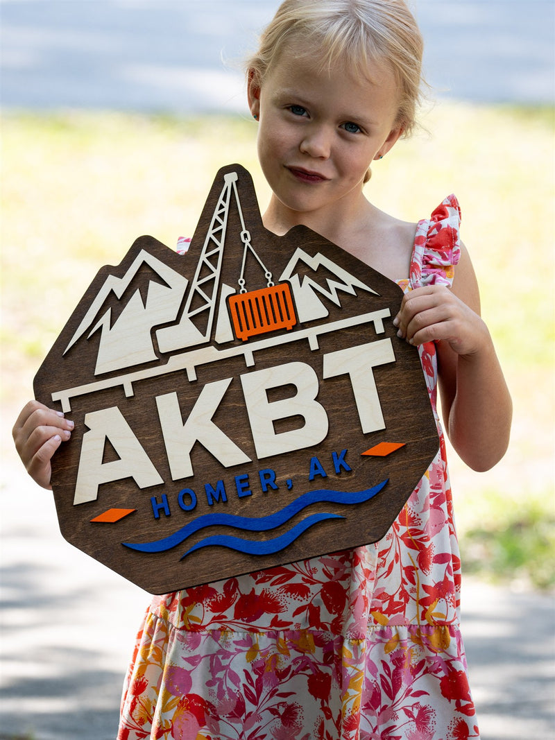 Mountain-shaped dark walnut logo sign for AKBT of Homer Alaska with a birch lattice crane lifting an orange shipping container