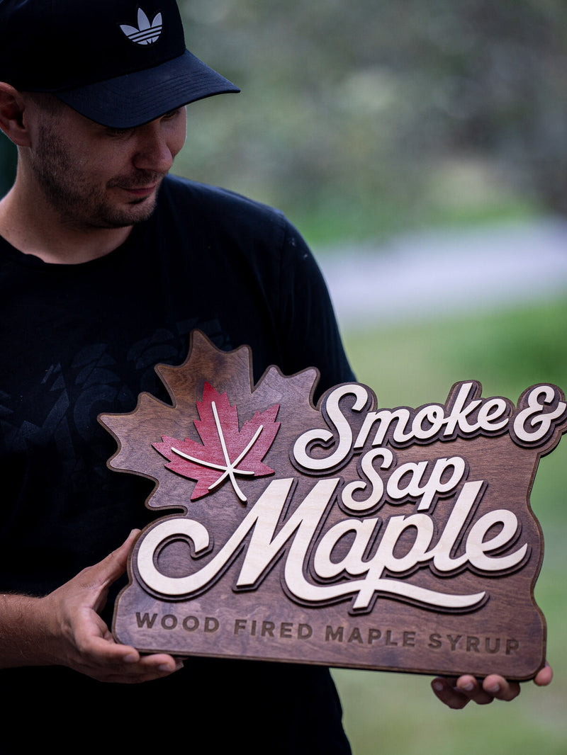 Shaped walnut sign with a maple leaf silhouette cut into the left edge holding a red stained leaf, reading Smoke and Sap Maple, wood fired maple syrup